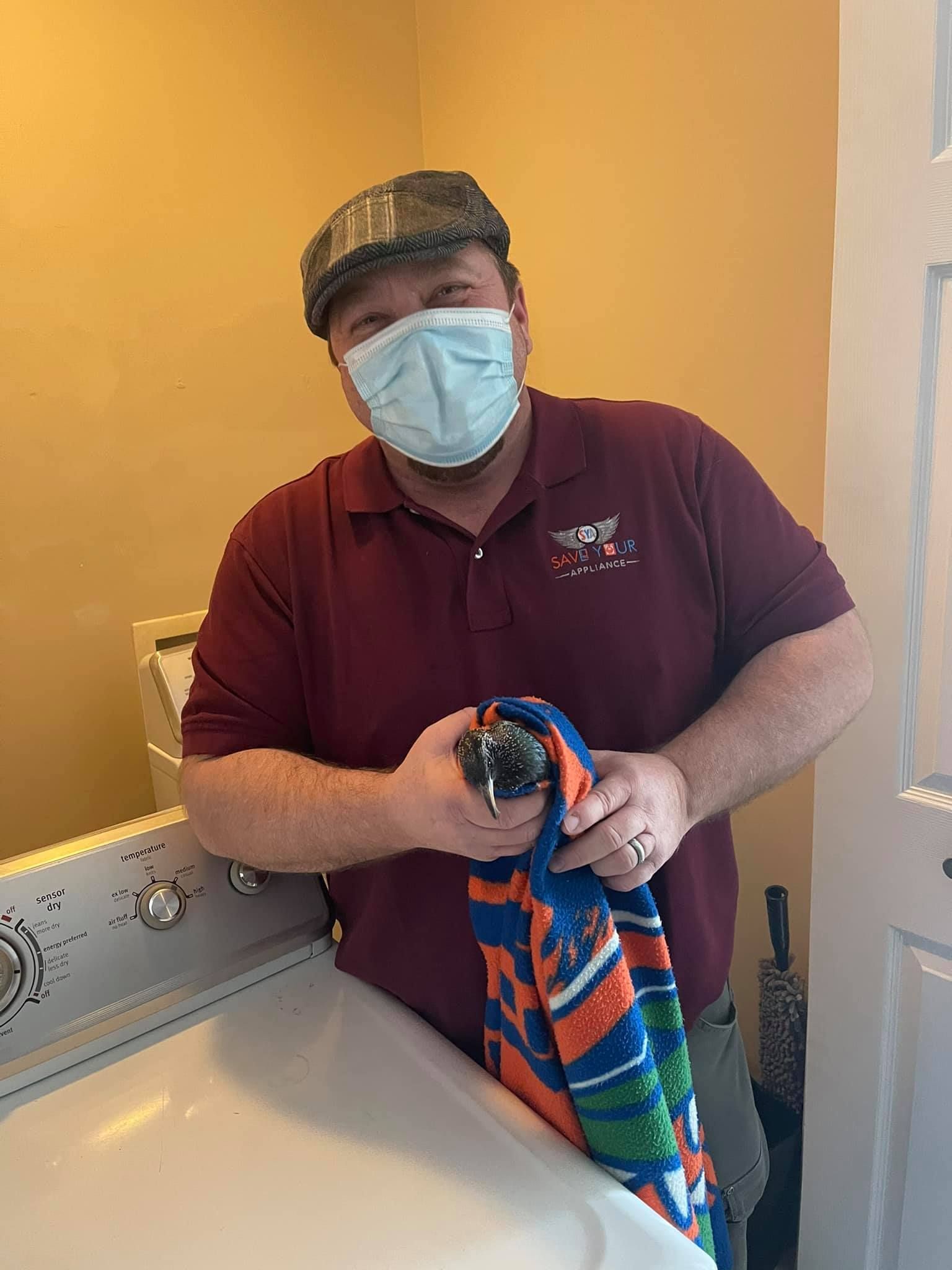 a man wearing a mask is standing next to a washing machine holding a towel .
