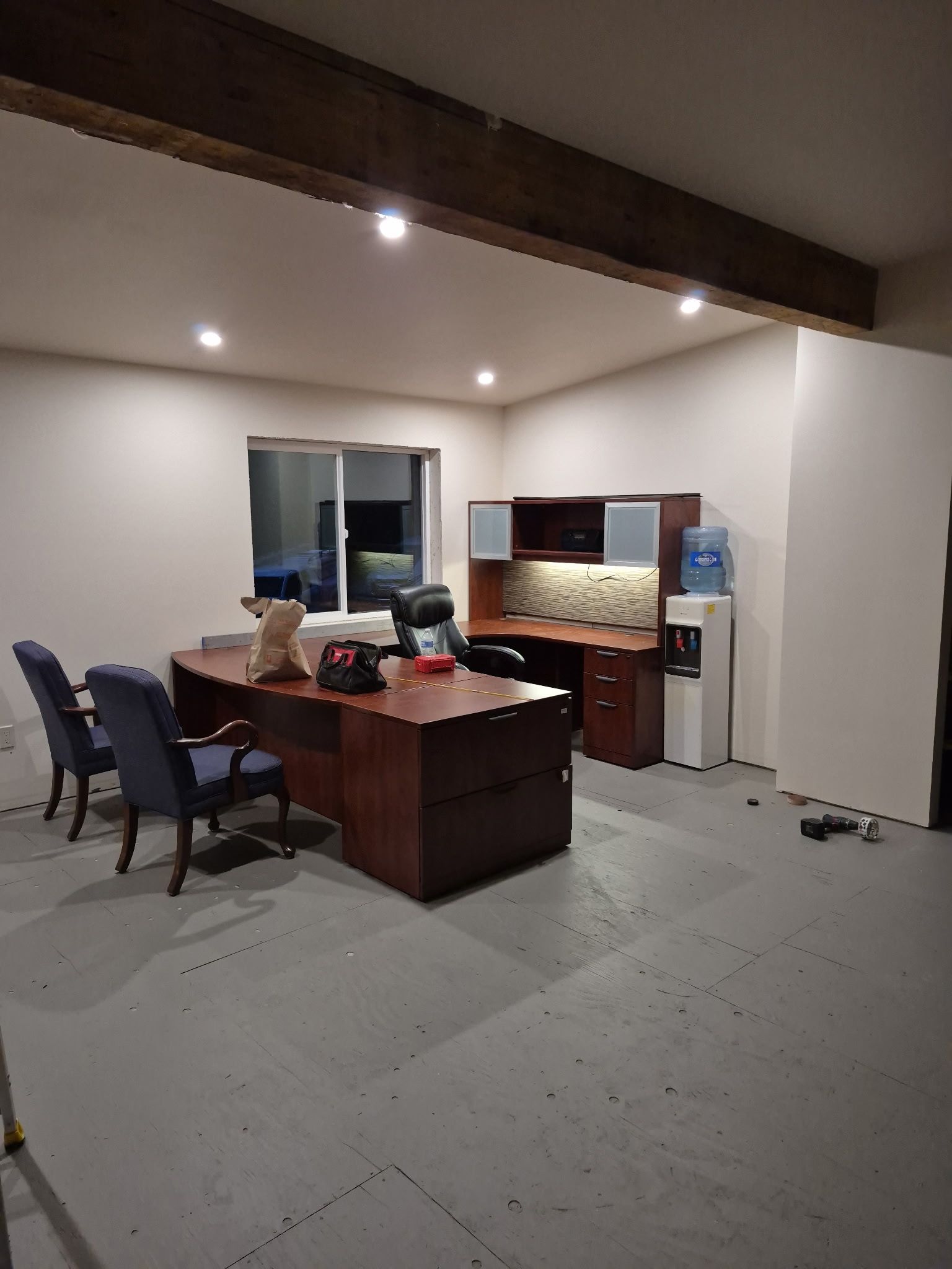 Office space with a large desk, chairs, and water cooler. Dark wooden beams and a window.