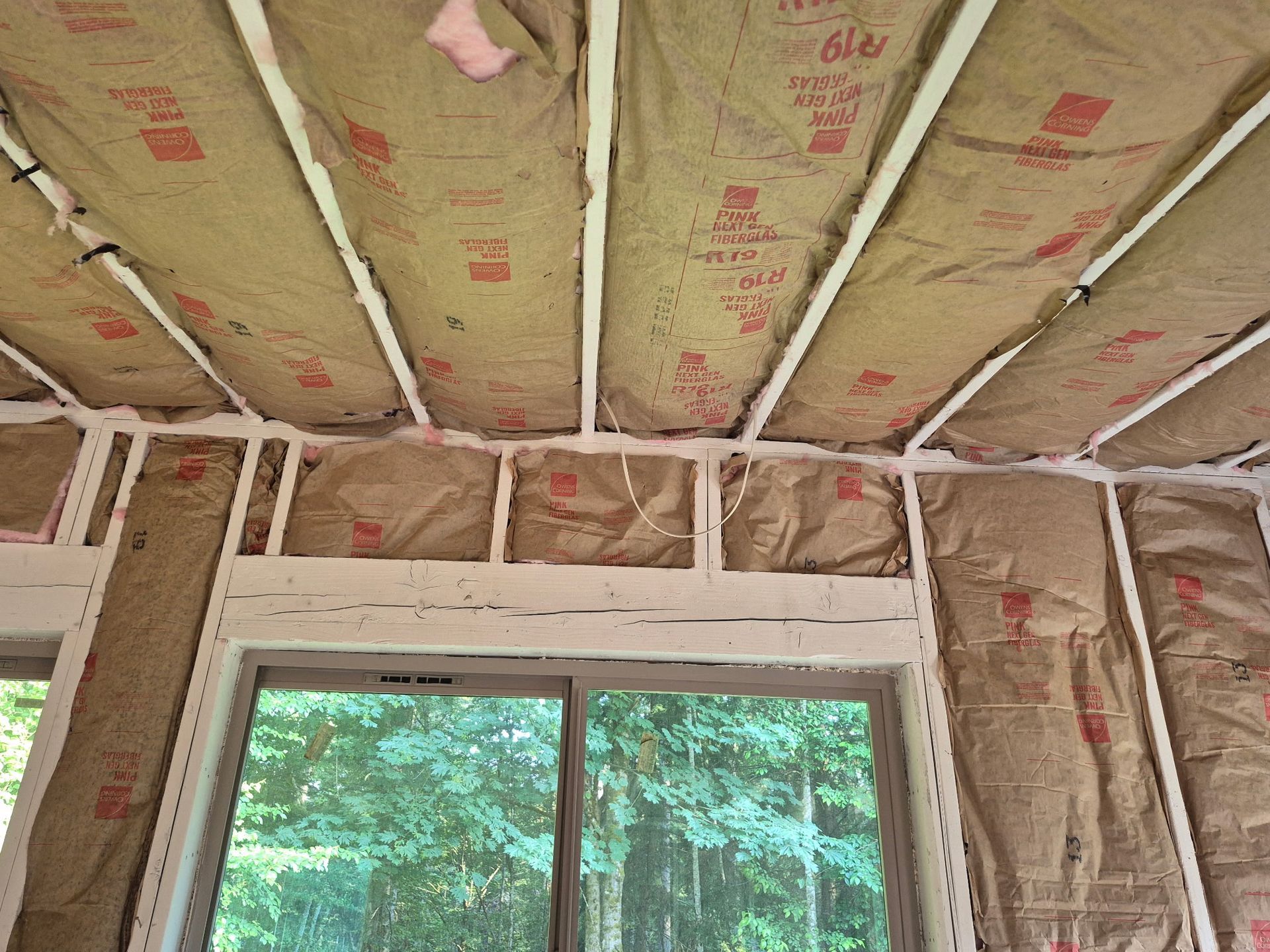 Insulation installed in ceiling and walls of a room with a window, against a view of greenery.