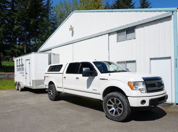 White pickup truck towing a white trailer parked in front of a white building on a sunny day.