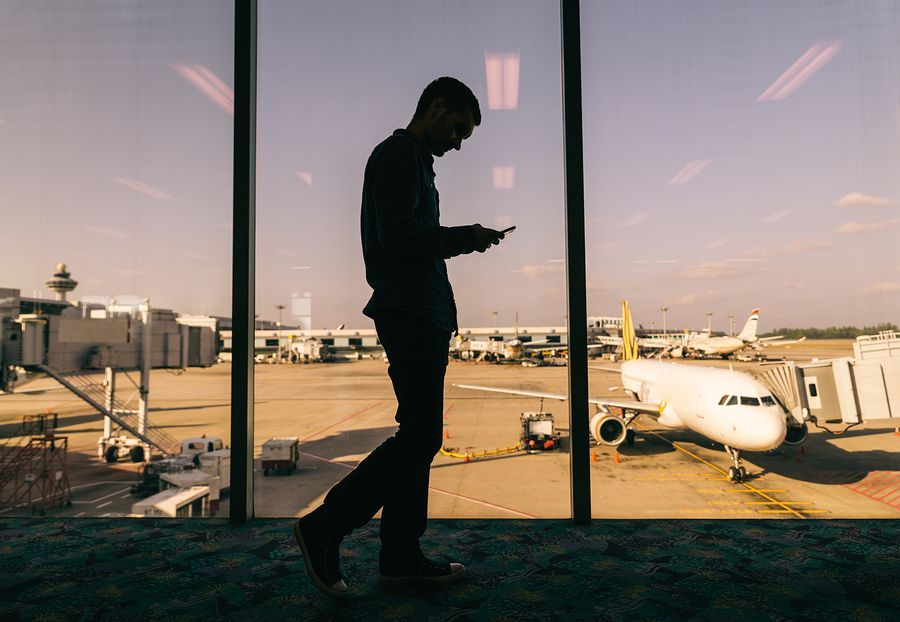 Silhouette of person at airport window, looking at phone. Airplanes and tarmac visible outside.