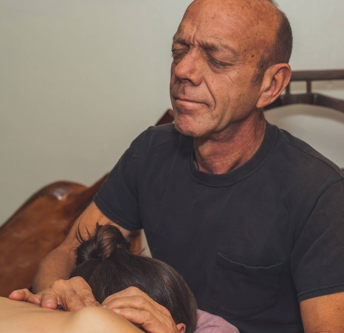 Man massaging another person's back, eyes closed. Indoors, neutral colors.