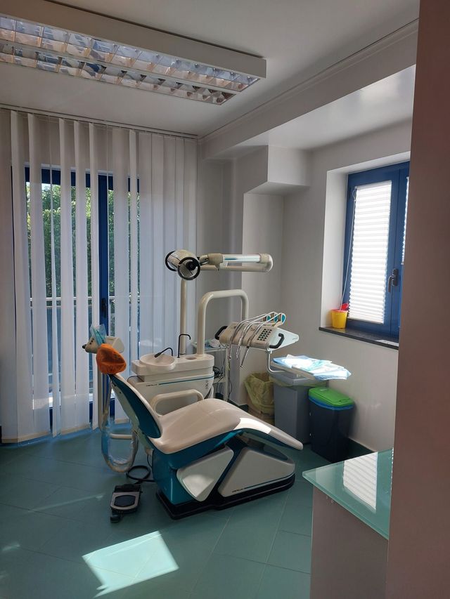 Sala per visite odontoiatriche con poltrona, lampada a soffitto e tende blu alla finestra.