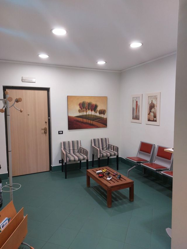 Piccola sala d'attesa con pavimento verde, porta in legno, sedie, tavolino e opere d'arte alle pareti.