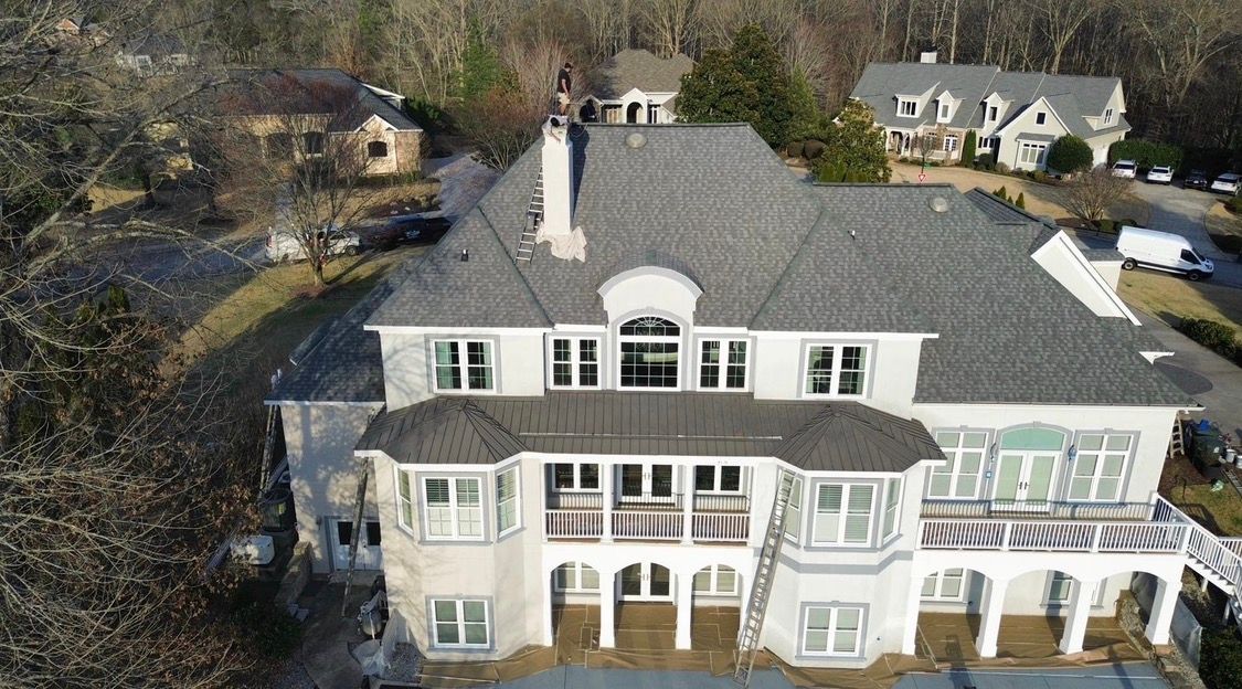 Aerial view of a large gray-and-white mansion with a front balcony and surrounding trees.