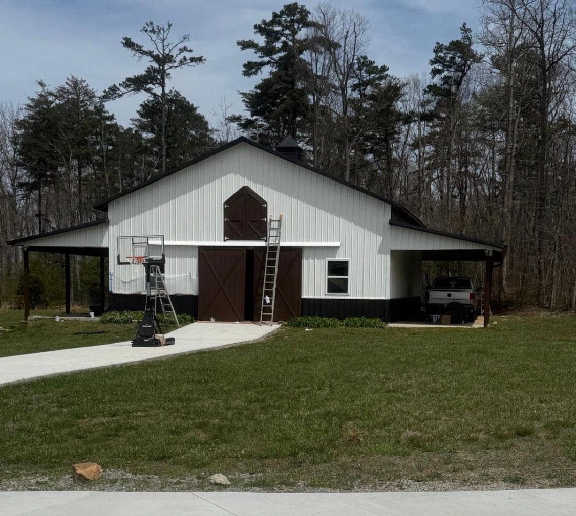 White barn-like house with a front walkway, small basketball hoop, and wooded background