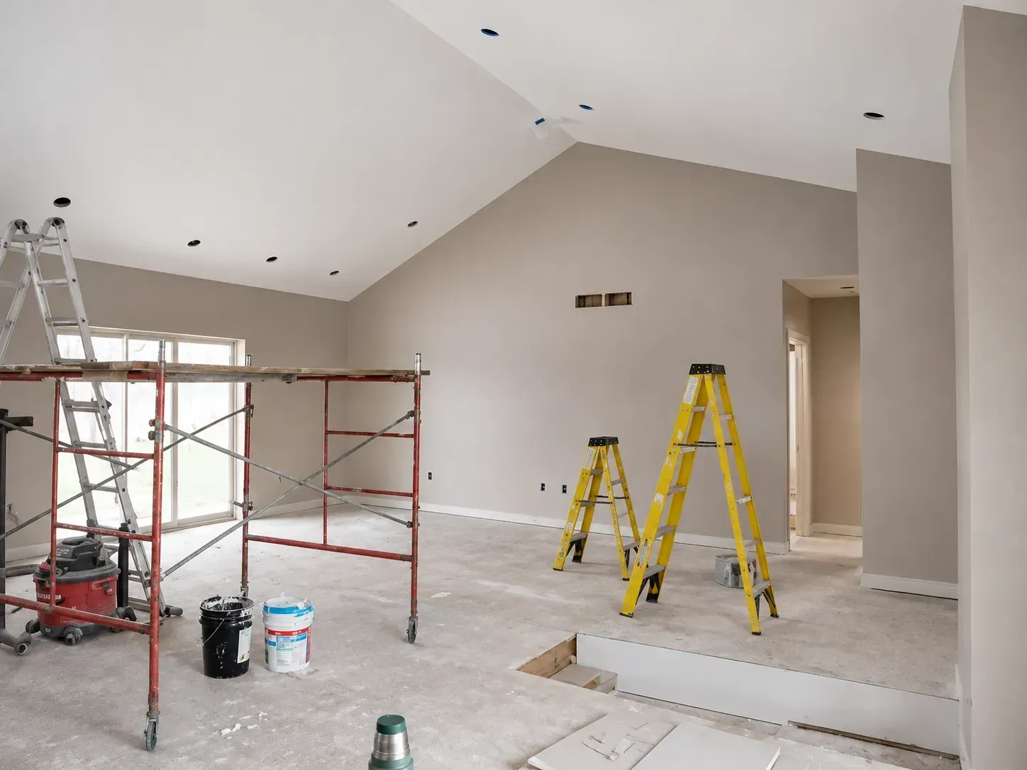 Partially renovated room with red scaffolding, yellow step ladders, and paint buckets under a high vaulted ceiling