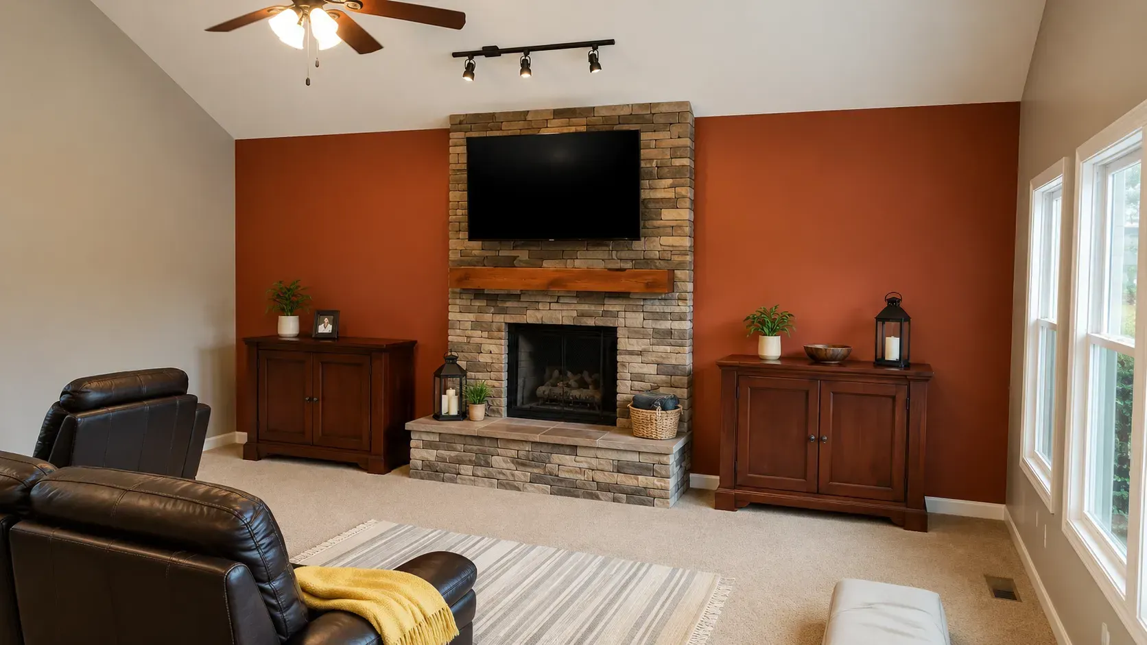 Living room with stone fireplace, wall-mounted TV, recliners, and large windows with beige walls.