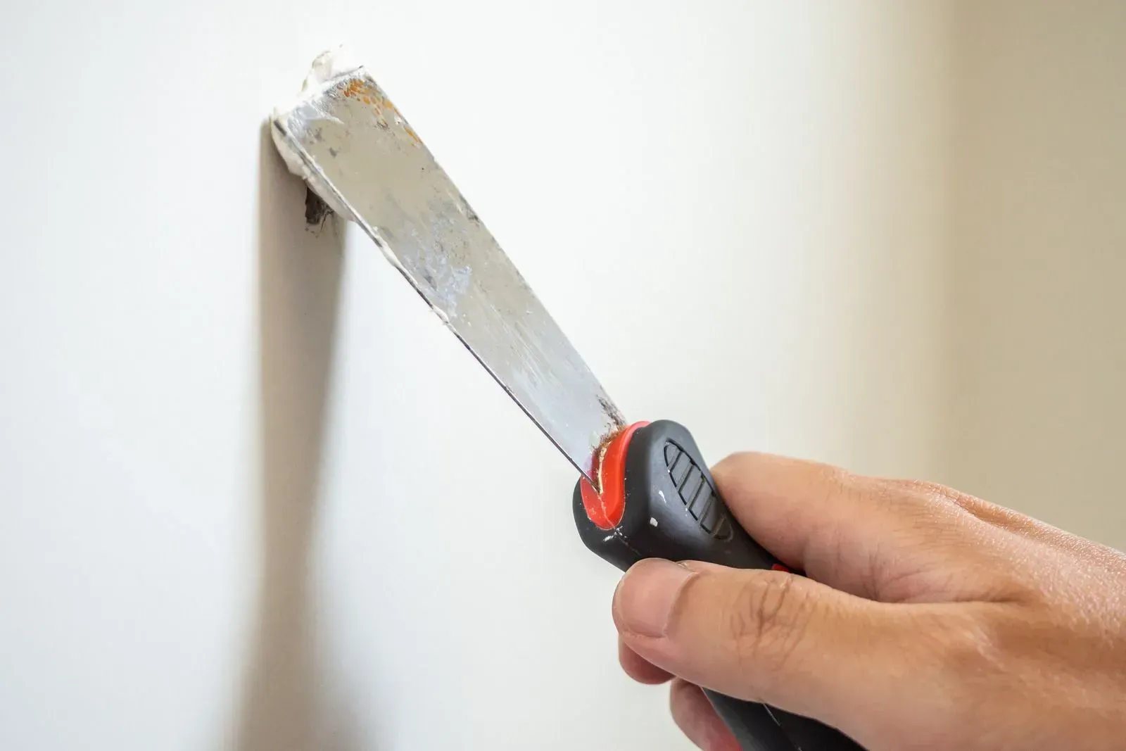 Hand using a putty knife to scrape paint from a white wall, creating a smooth edge
