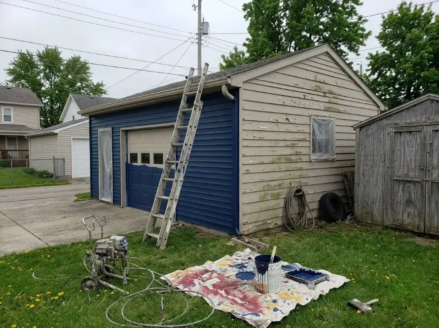 Blue garage with ladder leaning on it, paint supplies on tarp, and a weathered shed in a yard