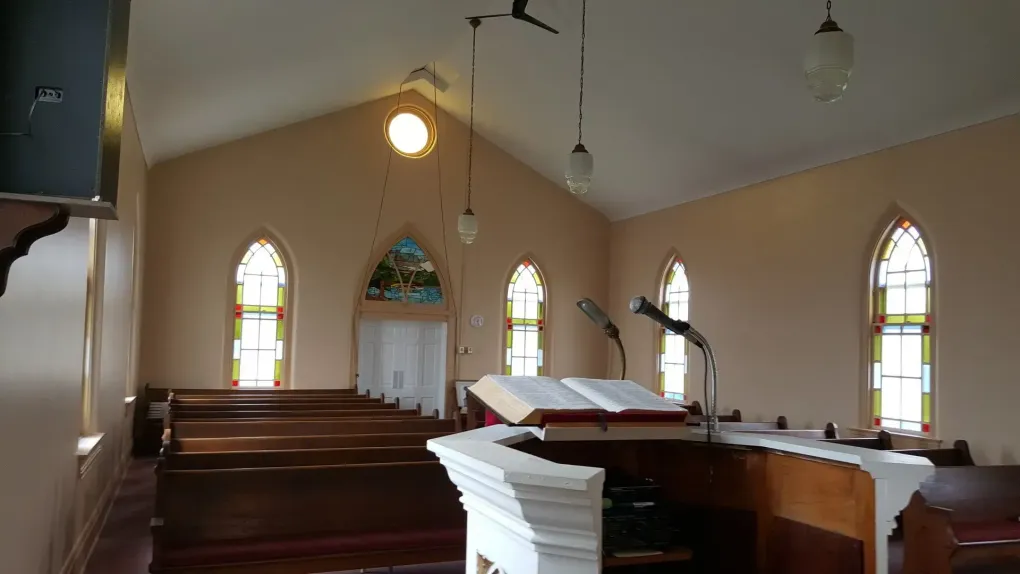 Interior of a small church sanctuary with wooden pews, stained-glass windows, and a pulpit at the front.