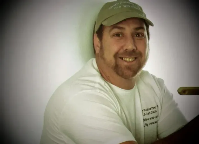 Smiling person in a white shirt and green cap sitting against a light background