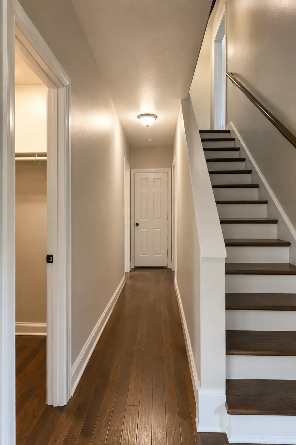 Narrow hallway with hardwood floors, white walls, ceiling light, and stairs on the right leading up