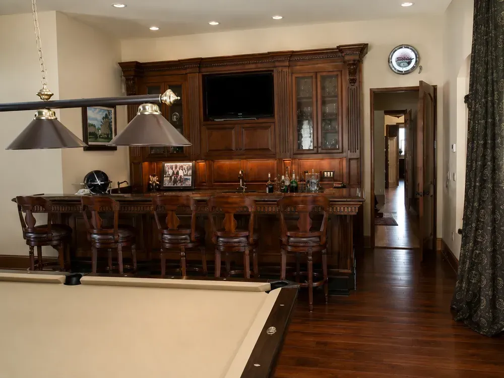 Warm wood-paneled home bar with stools, cabinets, and a pool table in the foreground