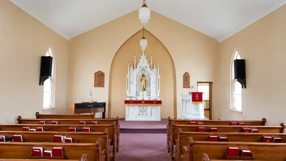 Interior of a small church sanctuary with wooden pews, red cushions, and a white altar at the front.