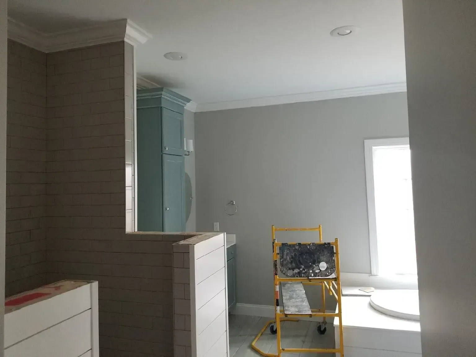 Unfinished room with gray walls, white cabinets, a brick column, and a yellow step ladder near a bright window.