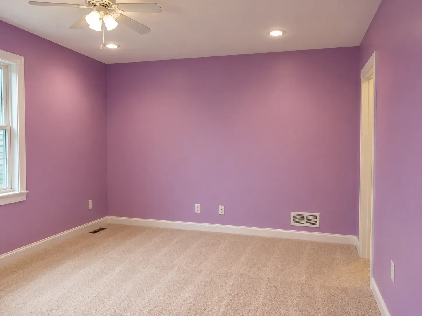 Empty lavender bedroom with beige carpet, white trim, ceiling fan, and windows on the left wall