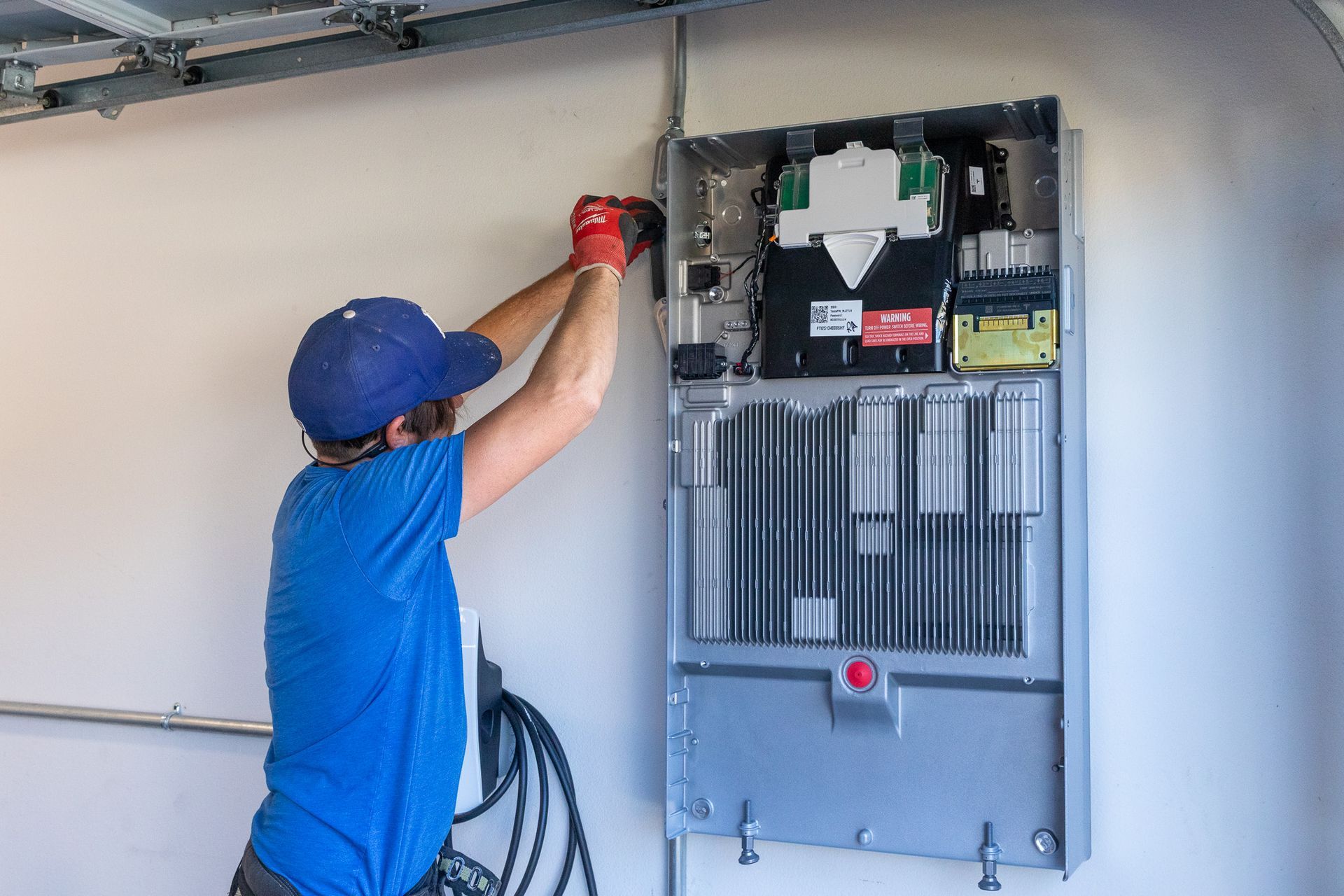 Worker in blue shirt and cap using a drill on an electrical panel in a garage.