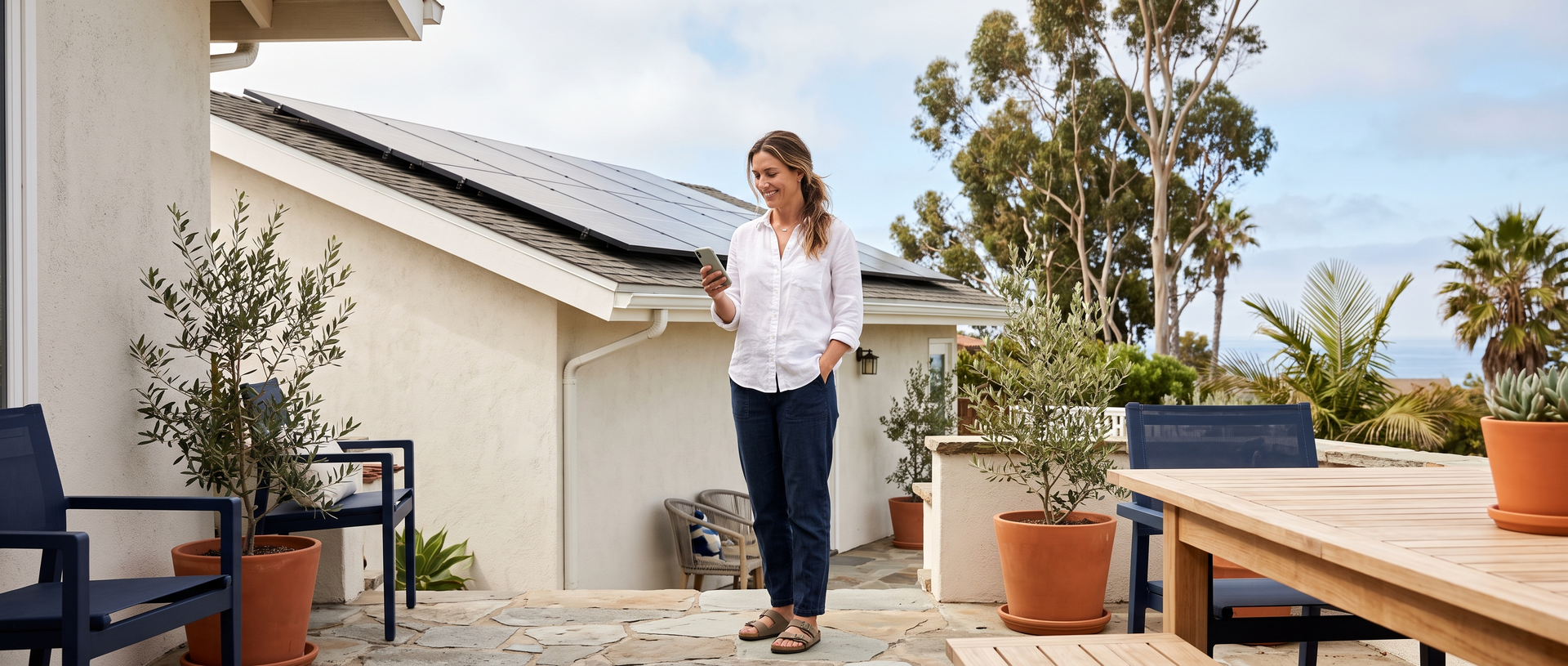 Homeowner checking a smartphone on the patio of a coastal home with rooftop solar panels
