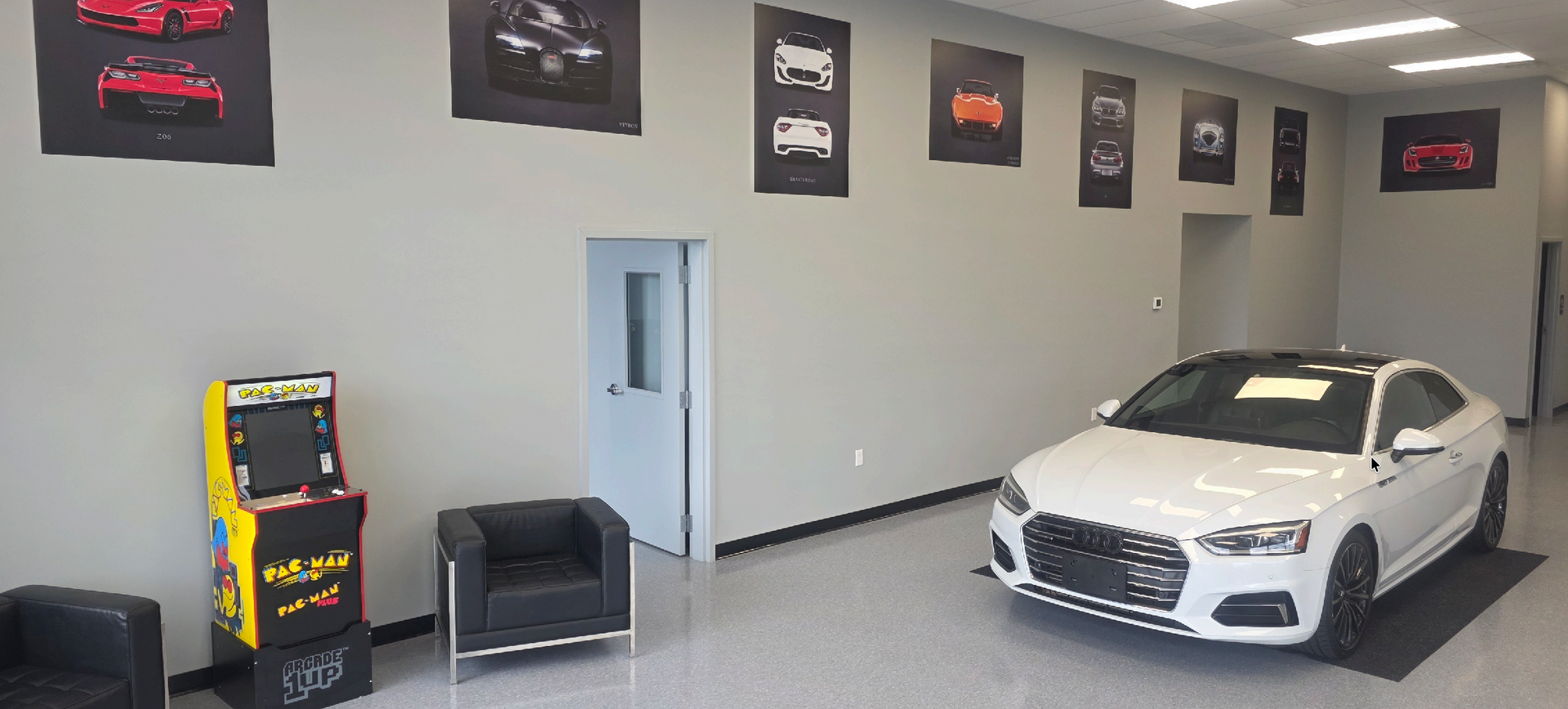 Car showroom with a white sedan, arcade machine, and wall posters in a clean, gray room. | Harford Auto Gallery