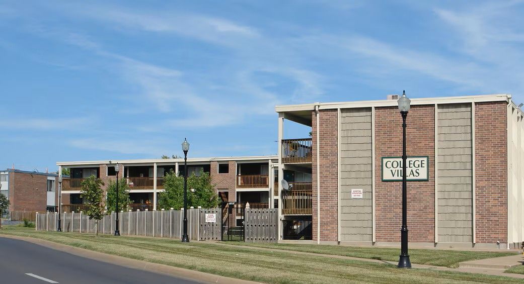 Two-story brick apartment building with balconies, wooden fence, and street sign under blue sky.