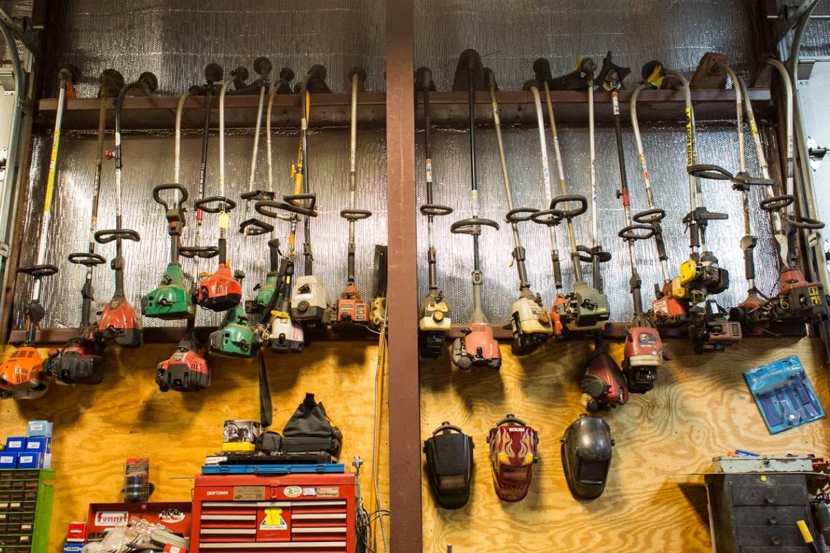 Row of gas-powered string trimmers hanging in a workshop, with red and black tool bags below.