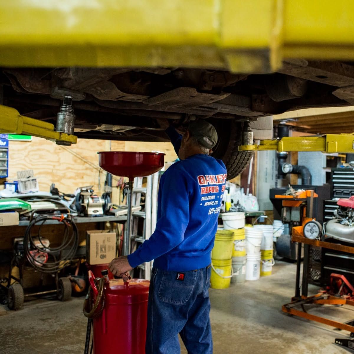 Mechanic in blue uniform under a lifted vehicle, draining red fluid in an auto repair shop