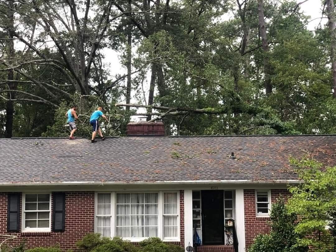 Two workers on a shingled roof beside a brick house with trees in the background
