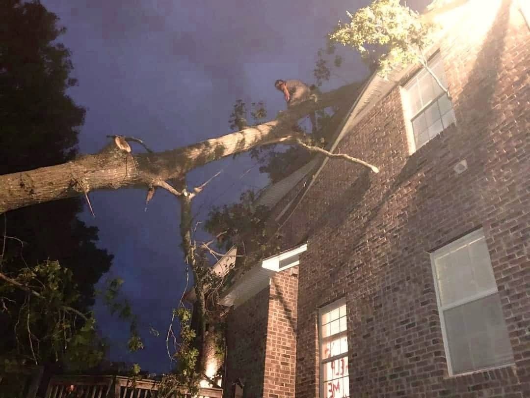 Large tree branch fallen onto a brick house at night, with a lit window and stormy sky