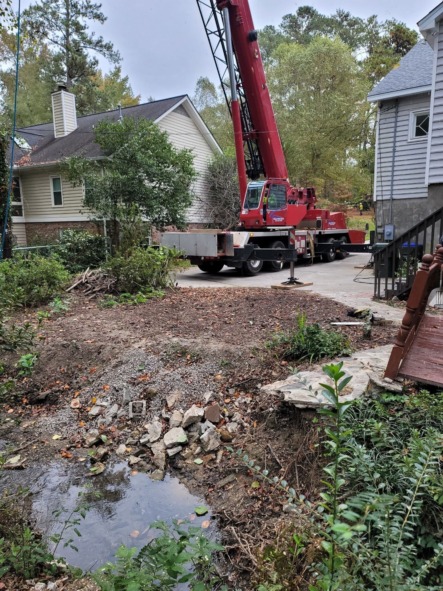Red crane truck in a wooded yard beside a house, with fallen leaves and a muddy puddle in the foreground.