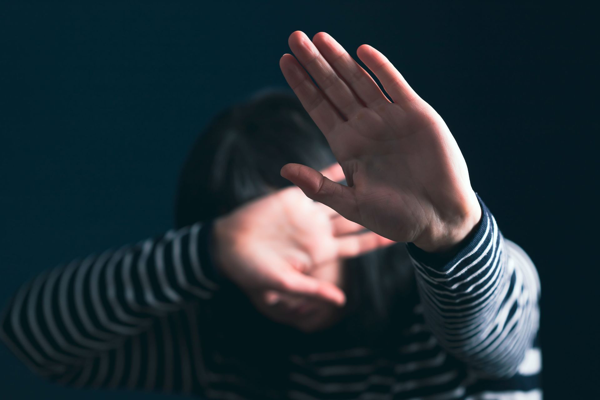 A person in a striped shirt holds their hands up in front of their face in a gesture of defense against a dark background.