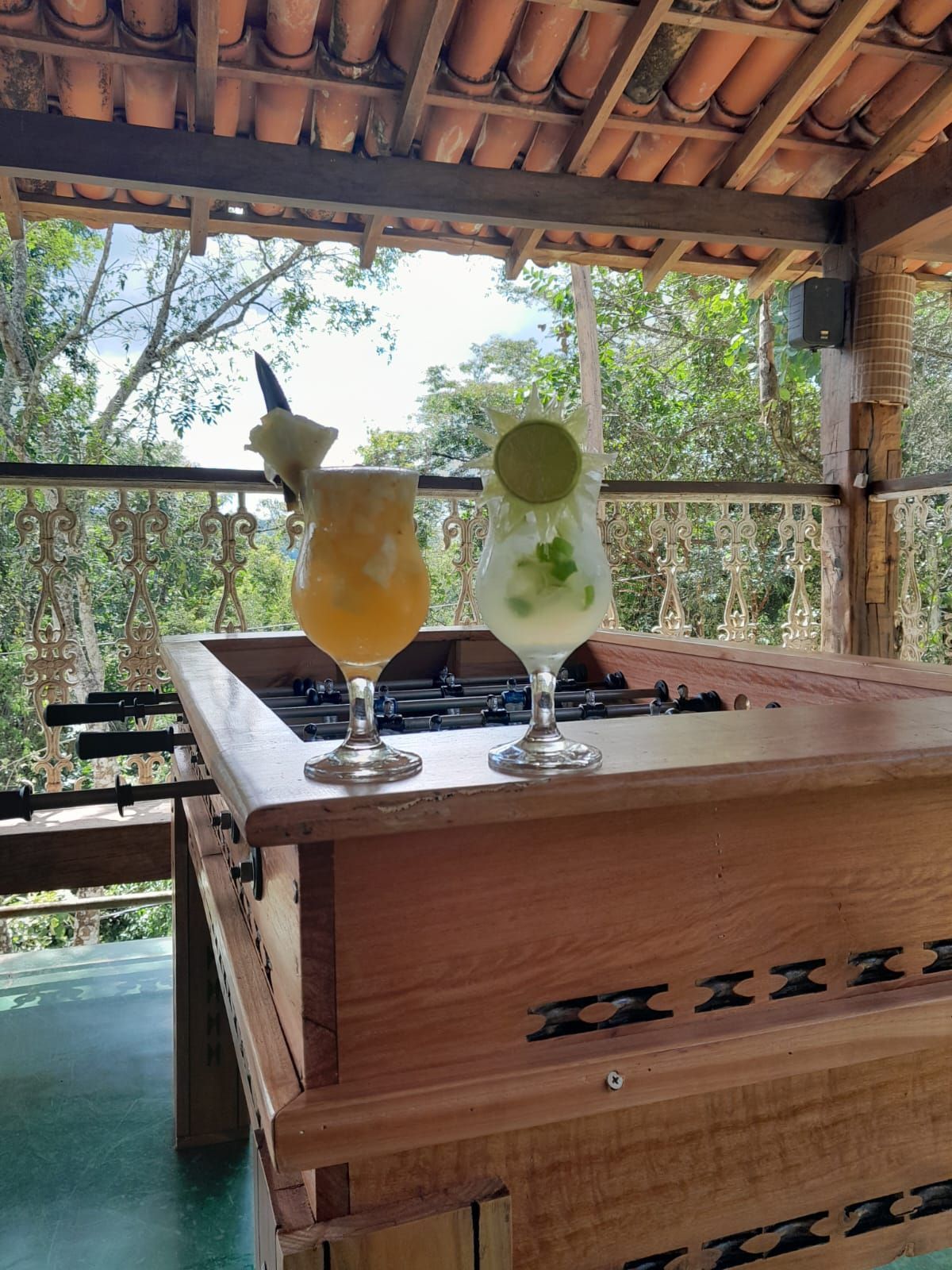 Dois coquetéis em uma mesa de pebolim numa varanda com vista para as árvores.