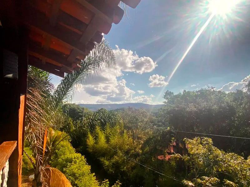 A luz do sol incide sobre uma paisagem florestal exuberante e ondulada, vista da varanda de madeira de uma casa sob um céu azul parcialmente nublado.