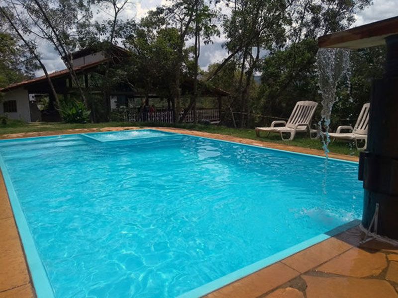 Uma piscina azul brilhante com uma pequena cascata, rodeada por um deck de pedra e duas espreguiçadeiras num jardim.