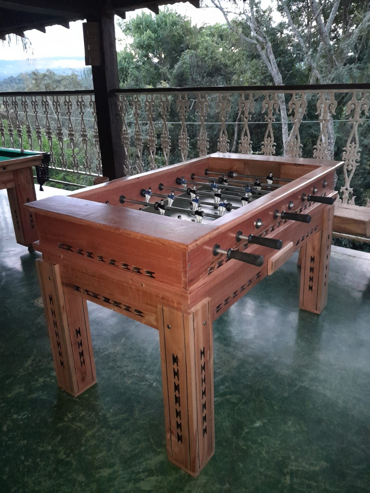 Uma mesa de matraquilhos de madeira num terraço com uma grade de metal e árvores ao fundo.