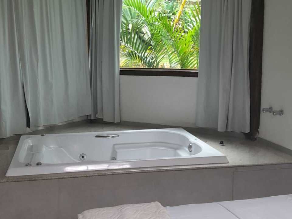 Uma banheira de hidromassagem branca instalada em uma plataforma ao lado de uma janela com cortinas brancas e vista para plantas verdes.