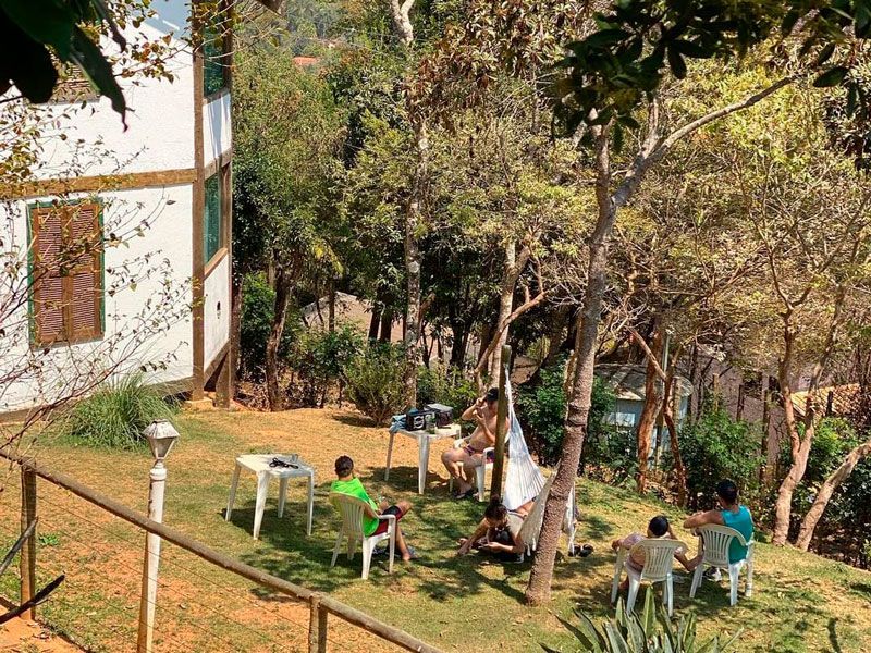 Pessoas sentam-se em cadeiras e relaxam em uma área gramada ensolarada e sombreada por árvores, ao lado de uma casa branca.