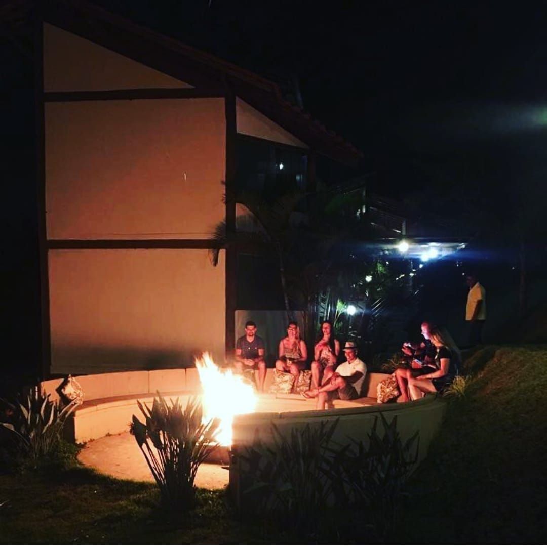 Um grupo de pessoas está sentado ao redor de uma fogueira acesa à noite, em frente a uma casa, desfrutando de um encontro social acolhedor.
