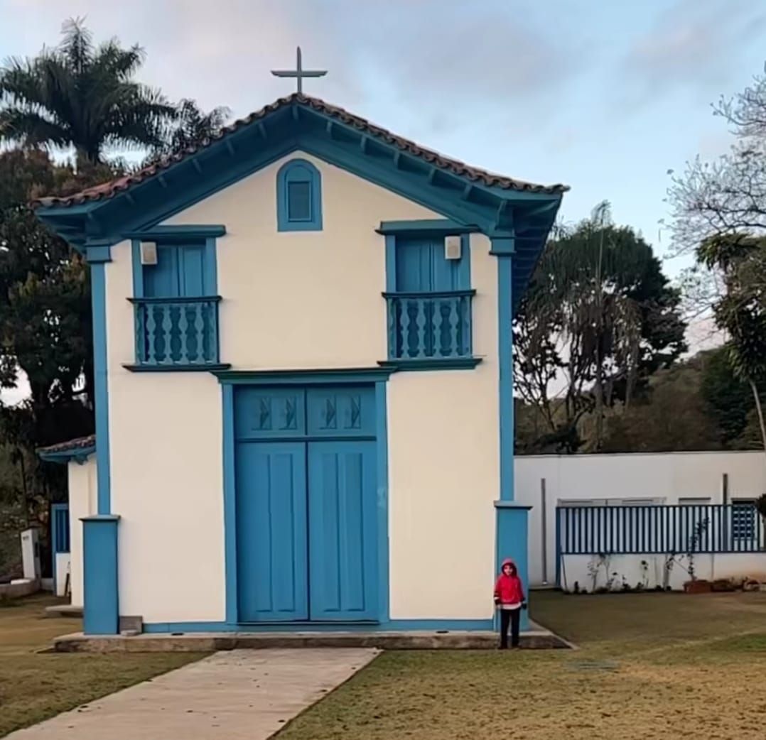 Uma pequena igreja branca com detalhes em azul e uma cruz no telhado, apresentando uma porta alta e uma pessoa em pé na frente.
