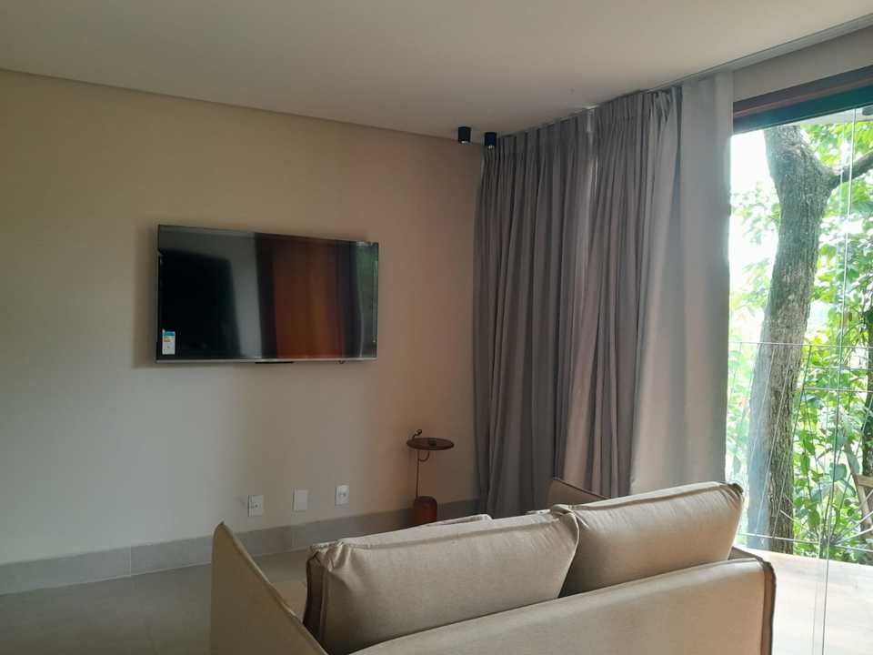 Uma sala de estar moderna com TV fixada na parede, sofá bege e cortinas cinza do chão ao teto ao lado de uma janela com vista para as árvores.
