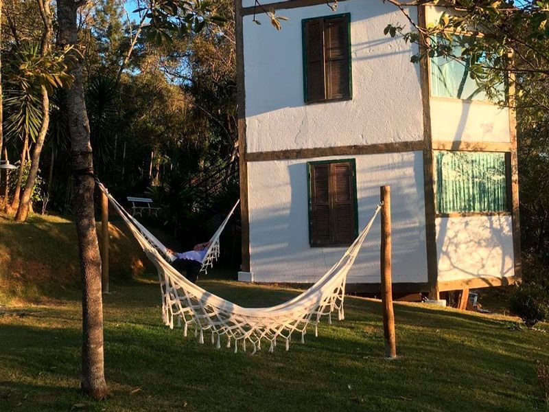 Uma casa branca de dois andares se ergue em um gramado, com uma pessoa descansando em uma rede branca estendida entre as árvores.