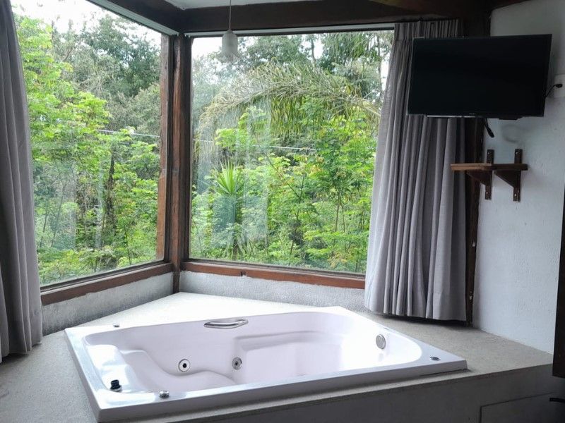 Uma banheira de canto branca fica ao lado de grandes janelas com vista para uma floresta verdejante e exuberante, com uma televisão instalada nas proximidades.