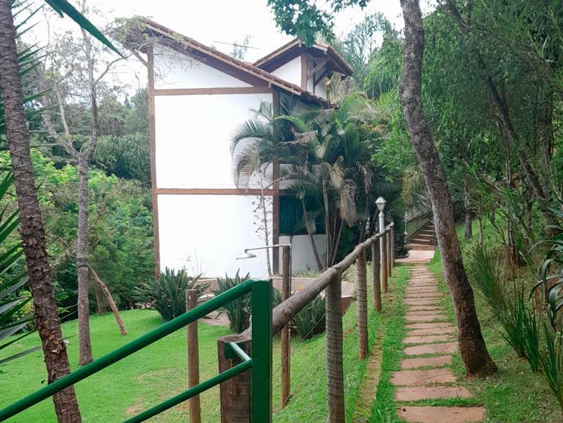 Uma casa branca com detalhes em madeira está situada em uma área arborizada e exuberante, ao lado de um caminho de pedra e uma cerca de metal verde.