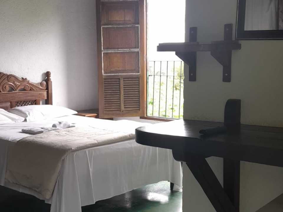 Um quarto com cama de madeira, lençóis brancos, uma estante e escrivaninha de madeira escura e uma porta de varanda aberta com vista para o exterior.