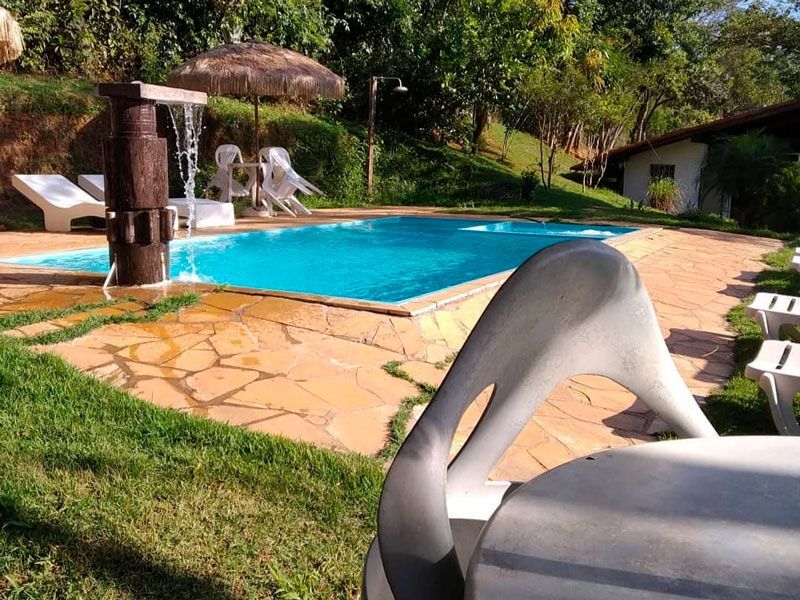 Uma piscina externa ensolarada com cascata, espreguiçadeiras e um pátio, emoldurada por uma área verde e arborizada.