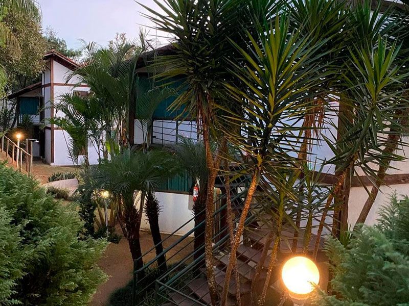 Um prédio branco de dois andares com varanda, rodeado por palmeiras verdejantes e iluminado por luzes esféricas externas.