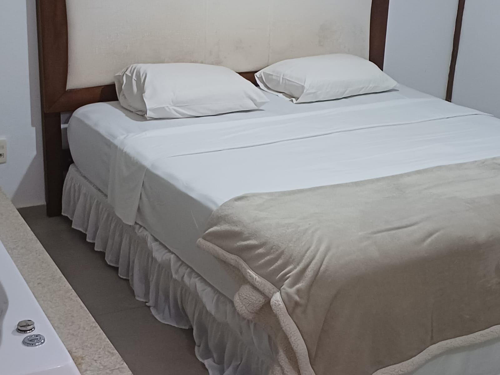 Uma cama arrumada com lençóis brancos e uma manta bege, com uma cabeceira de madeira escura encostada numa parede lisa.