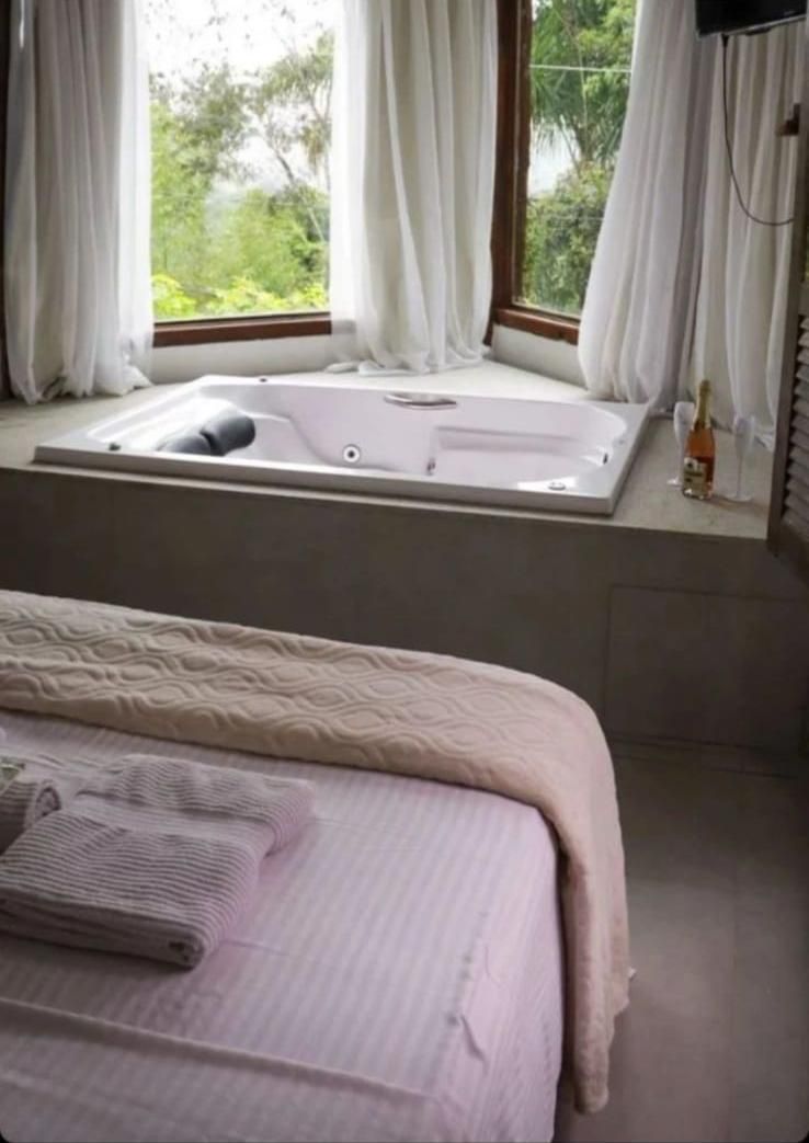 Um quarto com uma grande banheira de hidromassagem branca junto a uma janela com cortinas brancas, ao lado de uma cama com lençóis de cor clara.