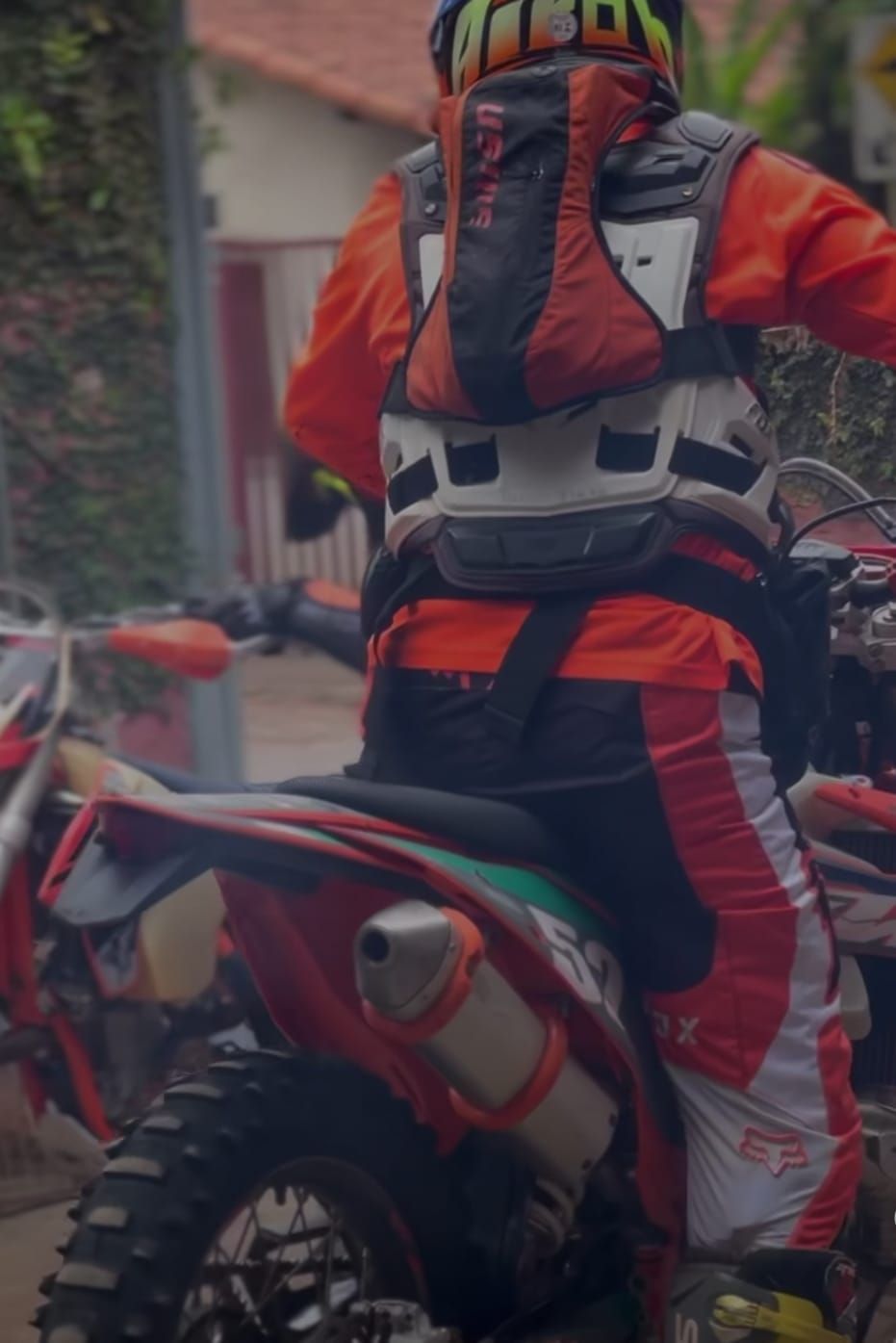 Um motociclista de motocross com equipamento laranja, preto e branco, incluindo um protetor de peito e uma mochila, sentado em uma motocicleta.