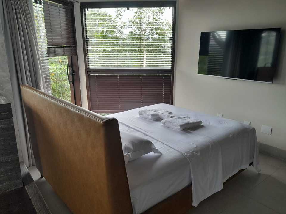 Um quarto limpo e moderno com cabeceira de couro, cama com roupa de cama branca e televisão fixada na parede em frente a uma grande janela.